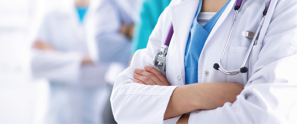 A close-up of a healthcare professional in a white lab coat with a stethoscope, standing with arms crossed. In the background, other healthcare professionals are slightly blurred, suggesting a team of doctors.