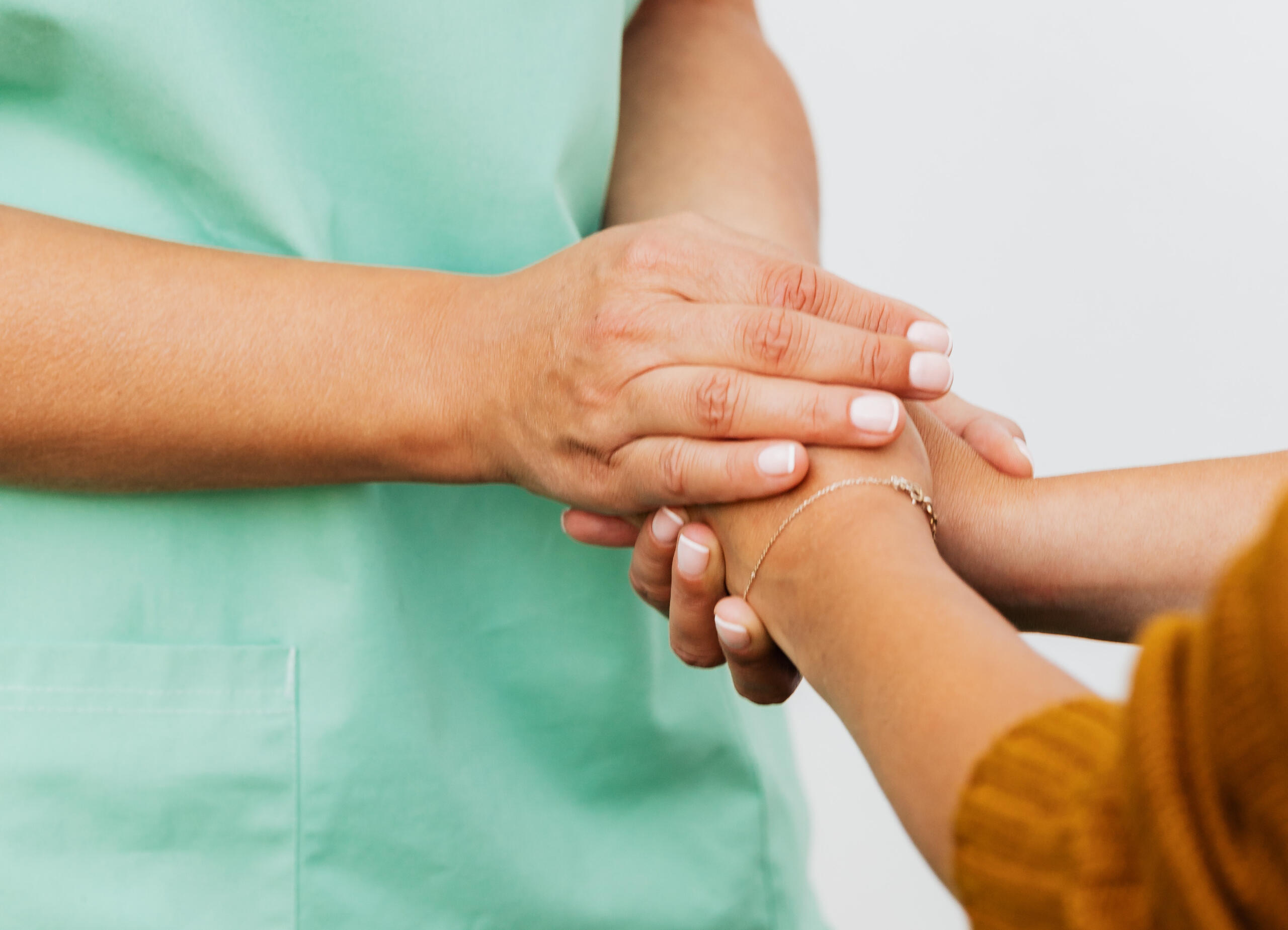 A healthcare professional in green scrubs gently holding the hand of a patient, offering comfort and support. The hands are clasped together, with a soft, reassuring gesture.