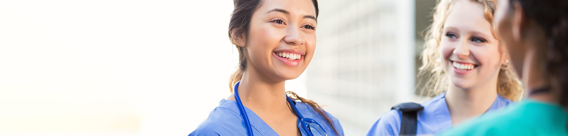 Three healthcare professionals in blue scrubs, smiling and engaging in a friendly conversation outdoors. One woman with a stethoscope is smiling at the camera, while the others appear to be smiling at her.