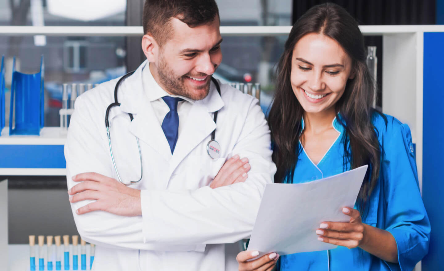 A smiling male doctor in a white coat and stethoscope, standing next to a female healthcare professional in a blue medical gown. They are both looking at a document in her hands, with a laboratory setting in the background.