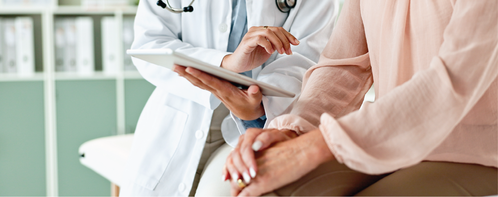 A healthcare professional in a white coat is discussing information on a tablet with a patient. The patient is seated, and the two are engaged in a conversation, with the healthcare professional pointing at the screen while the patient listens attentively.