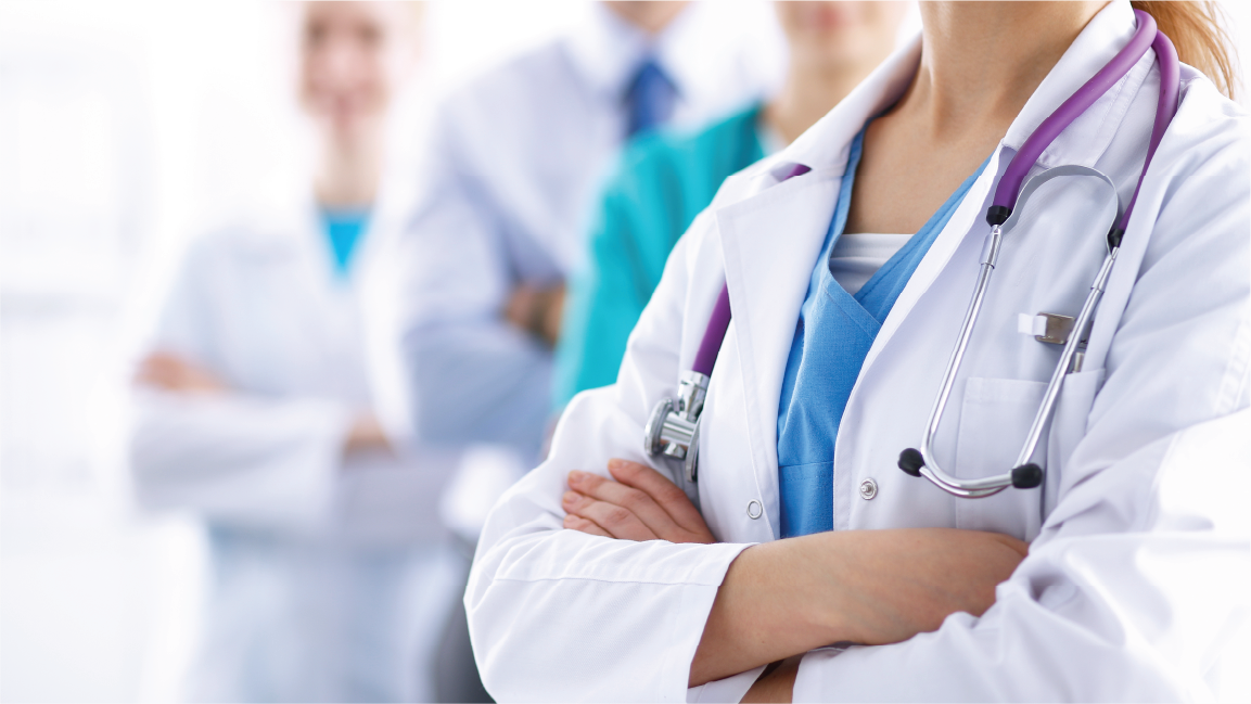A close-up of a healthcare professional in a white lab coat with a stethoscope, standing with arms crossed. In the background, other healthcare professionals are slightly blurred, suggesting a team of doctors.