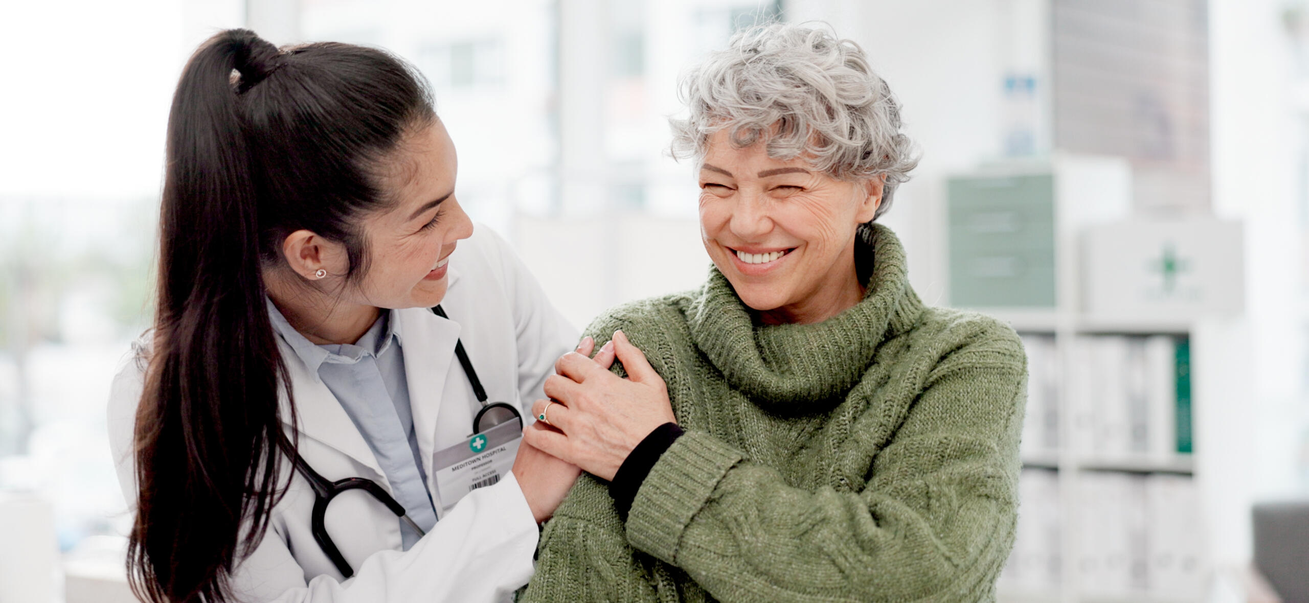 A healthcare professional in a white coat smiling and interacting with an elderly woman wearing a green sweater. The doctor has a hand on the patient's shoulder, and both are smiling warmly at each other in a caring, supportive environment.