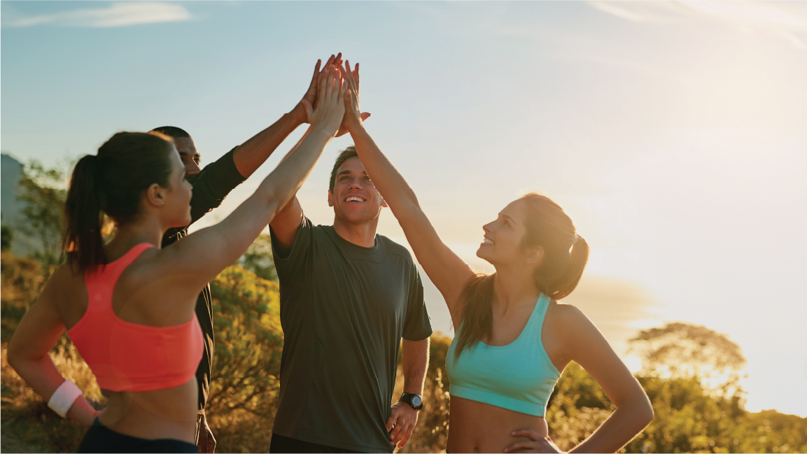 A group of four people, two women and two men, outdoors during sunset, giving each other high fives. They are dressed in activewear, smiling, and celebrating a moment of success or teamwork.
