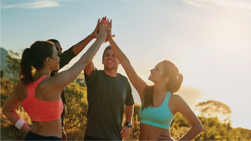A group of four people, two women and two men, outdoors during sunset, giving each other high fives. They are dressed in activewear, smiling, and celebrating a moment of success or teamwork.