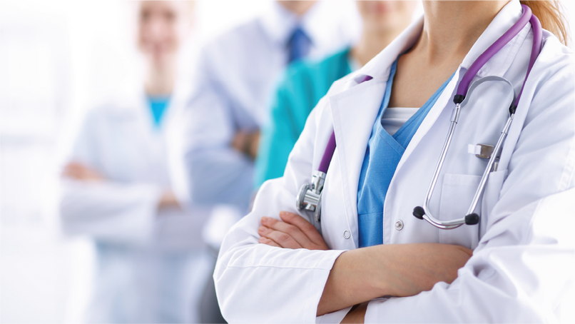 A close-up of a healthcare professional in a white lab coat with a stethoscope, standing with arms crossed. In the background, other healthcare professionals are slightly blurred, suggesting a team of doctors.